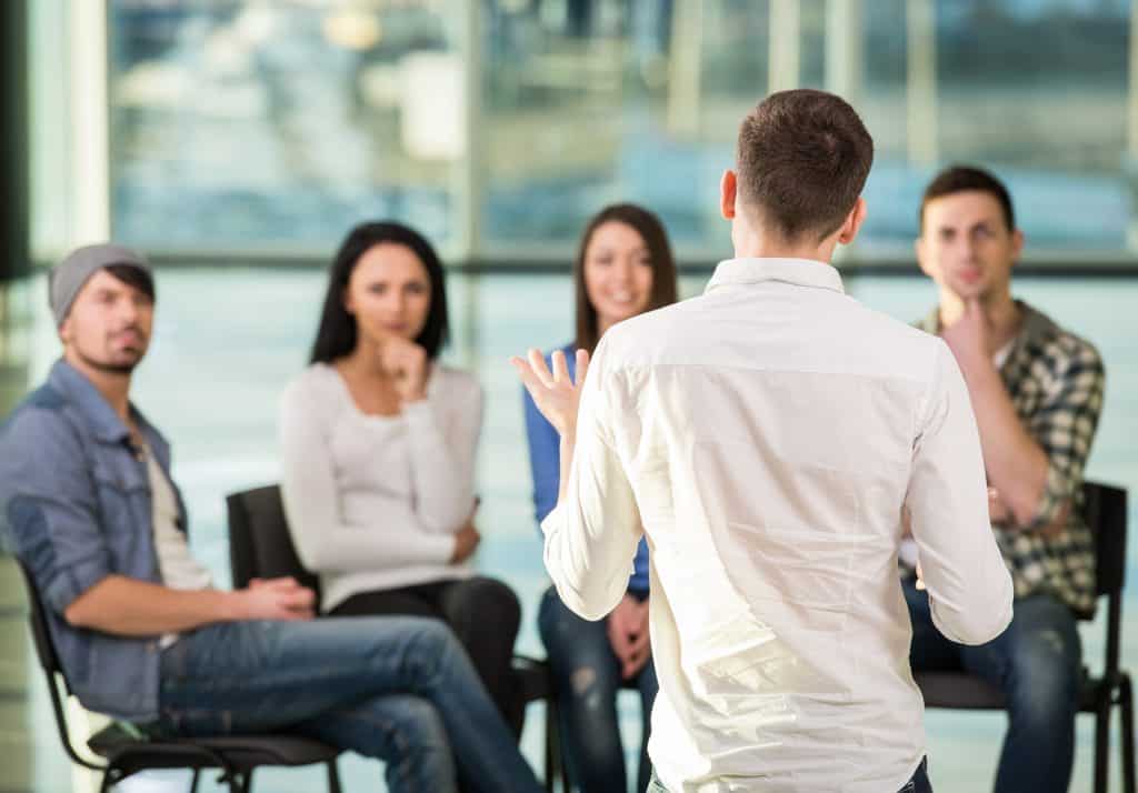 Young man sharing his problems with people. View of man is telling something and gesturing while group of people are sitting in front of him and listening.