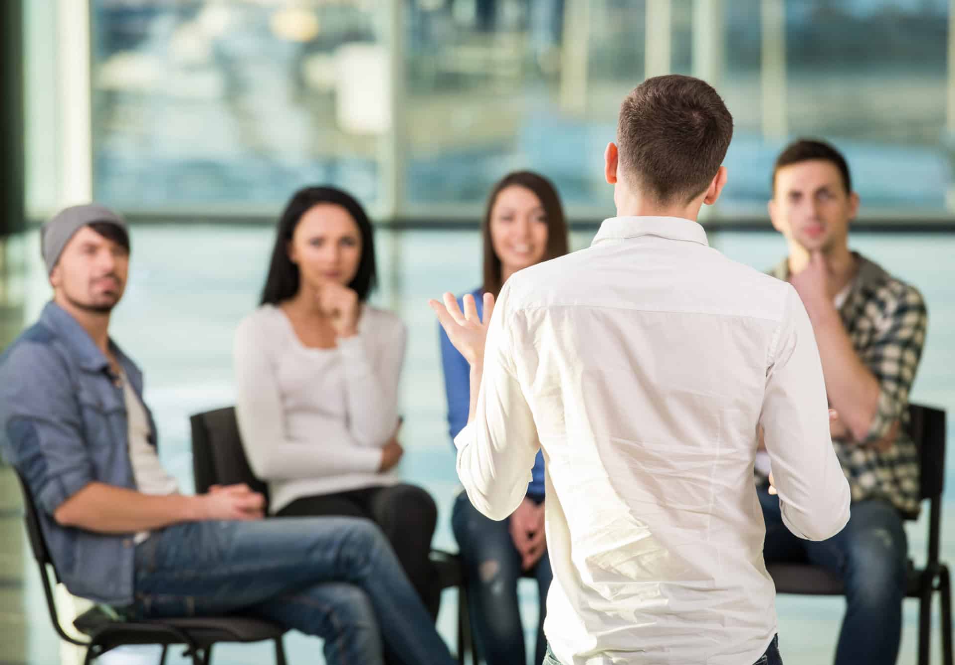 Young man sharing his problems with people. View of man is telling something and gesturing while group of people are sitting in front of him and listening.